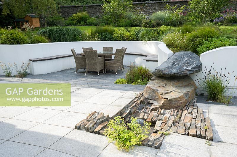 Contemporary terrace with rendered white retaining wall, naturalistic rock outcrop, dome fountain water feature and rattan garden furniture.