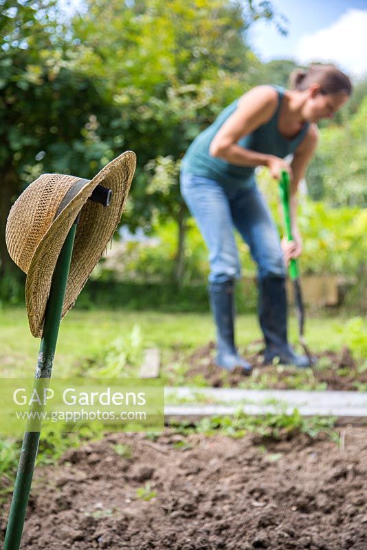 Woman digging and forking soil in an allotment patch. Focus on hat hung on edging knife