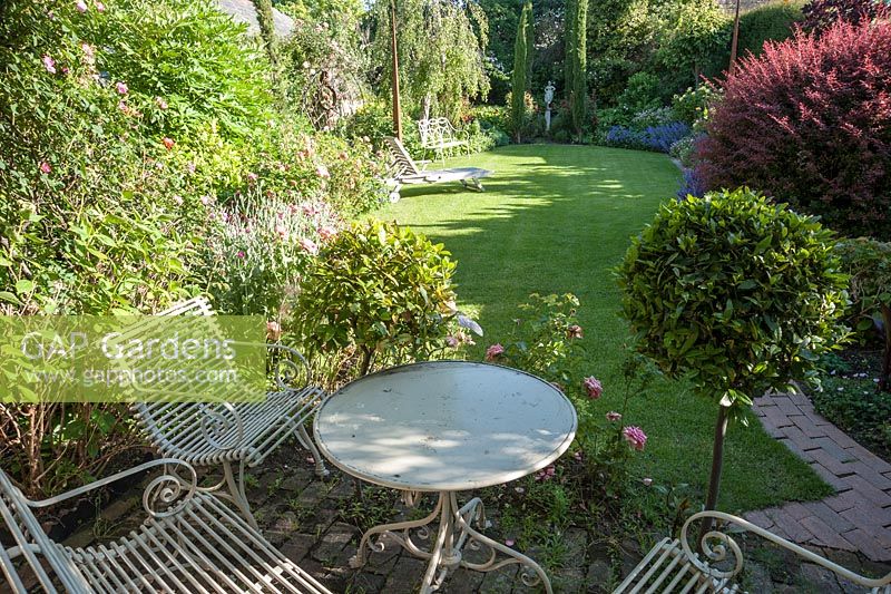 Seating area surrounded by clipped bays, with lawn, borders and pencil cypresses beyond. 