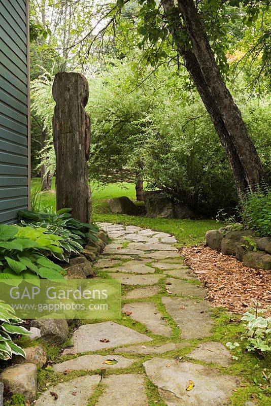 Jack-rabbit Johanssen wooden totem pole and flagstone path next to a border with Hostas in backyard garden in summer