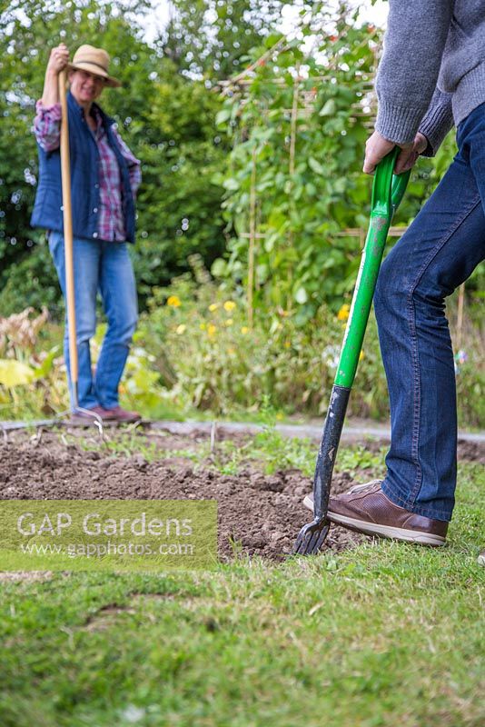 Man forking soil over in an alloment plot, woman standing watching in the background