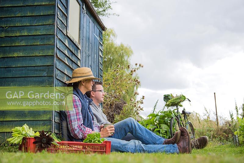 Man and woman sitting down against an allotment shed, crate of harvested produce in the foreground