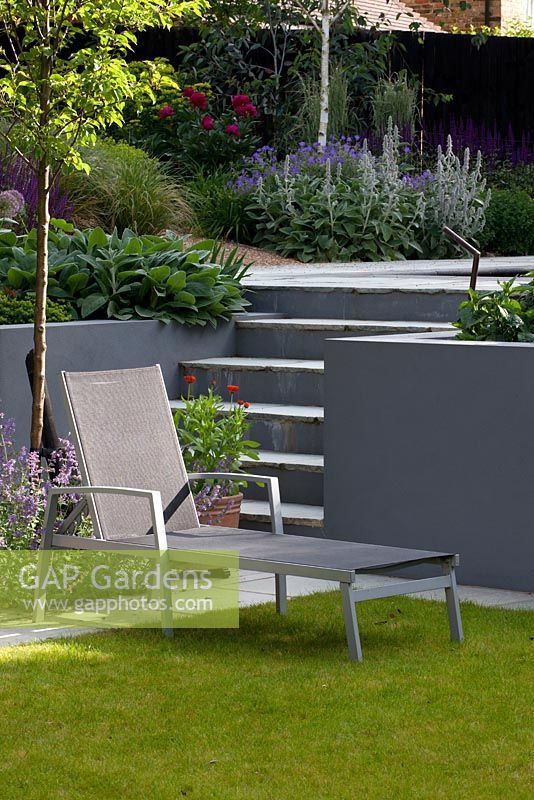 Sun lounger on small lawn next to grey painted walls and steps.  Planting includes Nepeta 'Six Hills Giant',