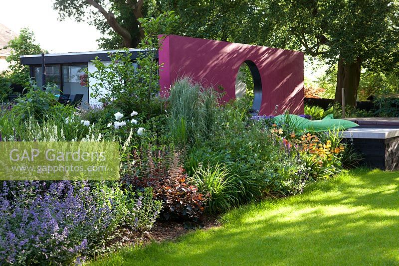 Modern garden with mixed flowerbeds, pink moon wall, raised wooden deck with bean bags in summer