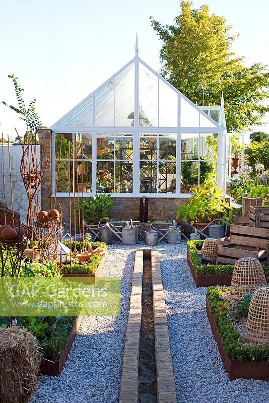 Greenhouse in garden, gravel path, metal framed raised beds and low box hedges, vegetables, strawberries and herbs planted in wooden crates, watering cans in background and water tiled basin with water tap, water feature with small canal
