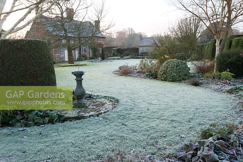 Frosty garden with mixed borders, clipped evergreens, white stemmed birches and a sundial with Elizabethan house beyond.