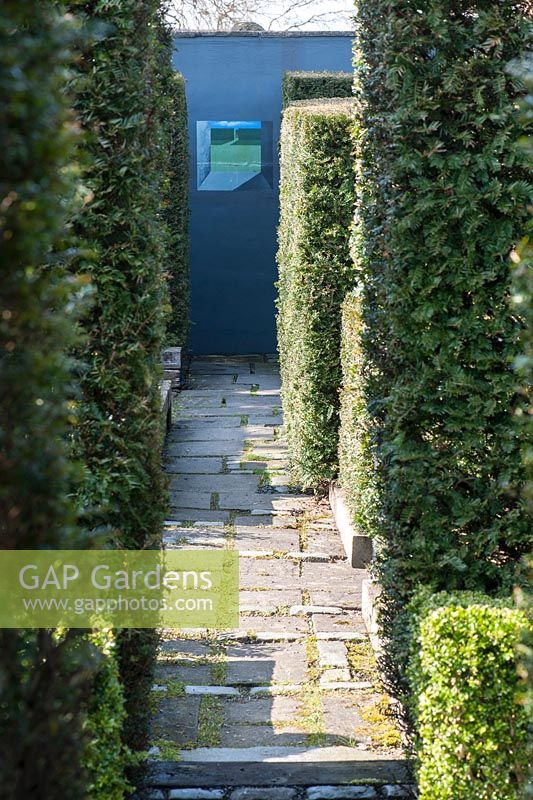 Yew hedges frame a view to a small painted image on a dark grey wall in a formal garden. 