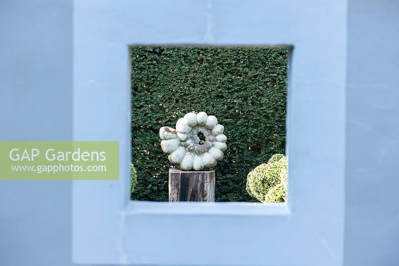 Ammonite sculpture by Darren Yeadon seen through an opening in a painted wall in a formal garden. 