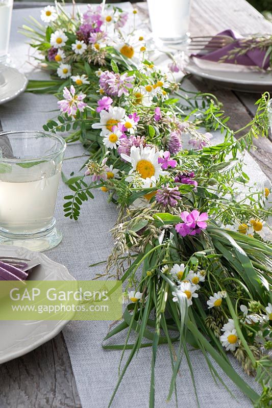 Early summer table decoration: table garland decorated with matricaria chamomilla - chamomile, leucanthemum vulgare - daisies, coronilla, trifolium pratense - red clover, dianthus and grasses