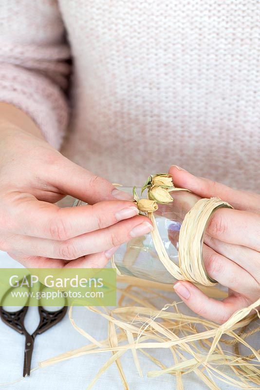 Decorating glass jars for garden posies step by step.  Attaching dried rose buds to raffia decoration on a glass jar.