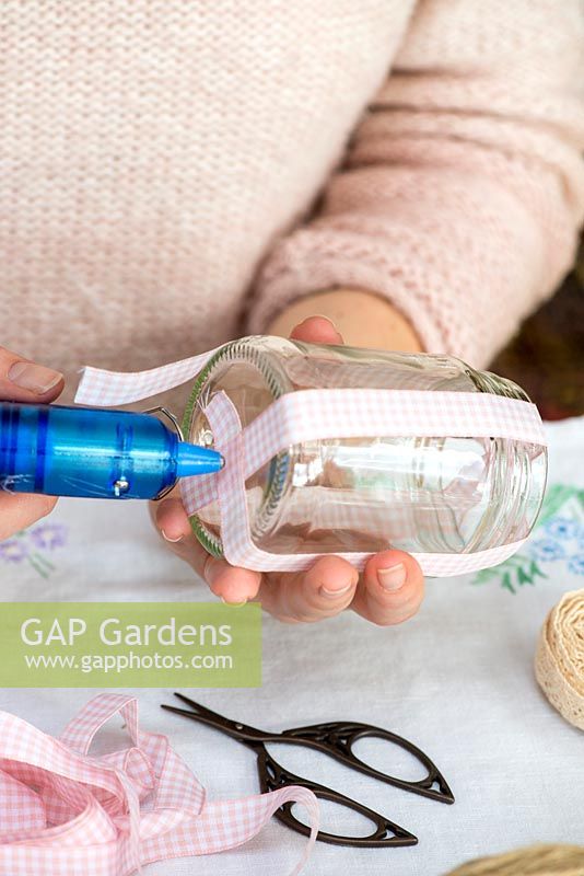 Decorating glass jars for garden posies step by step. Fixing a pink gingham ribbon to the base of a glass jar with a hot glue gun.