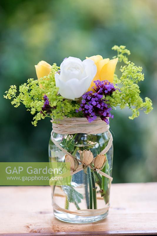 A summer posie with everyday flowers - Verbena bonariensis, Alchemilla mollis and yellow and white roses in glass jar decorated with raffia and dried rose buds.