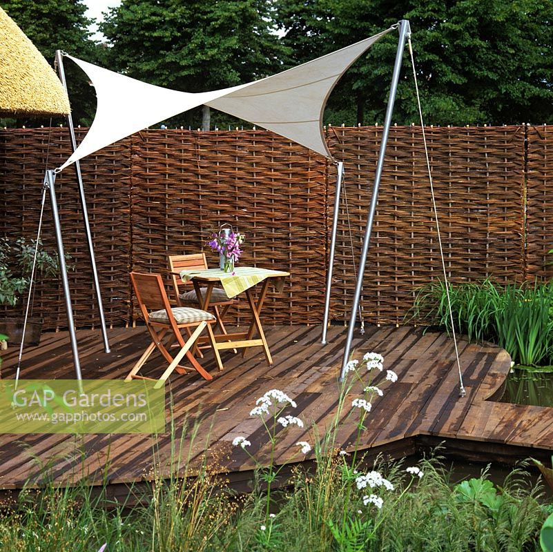 Canvas awning is strung, sail-like, between stainless steel posts to provide shade on a wooden deck. Privacy created from woven willow screens.