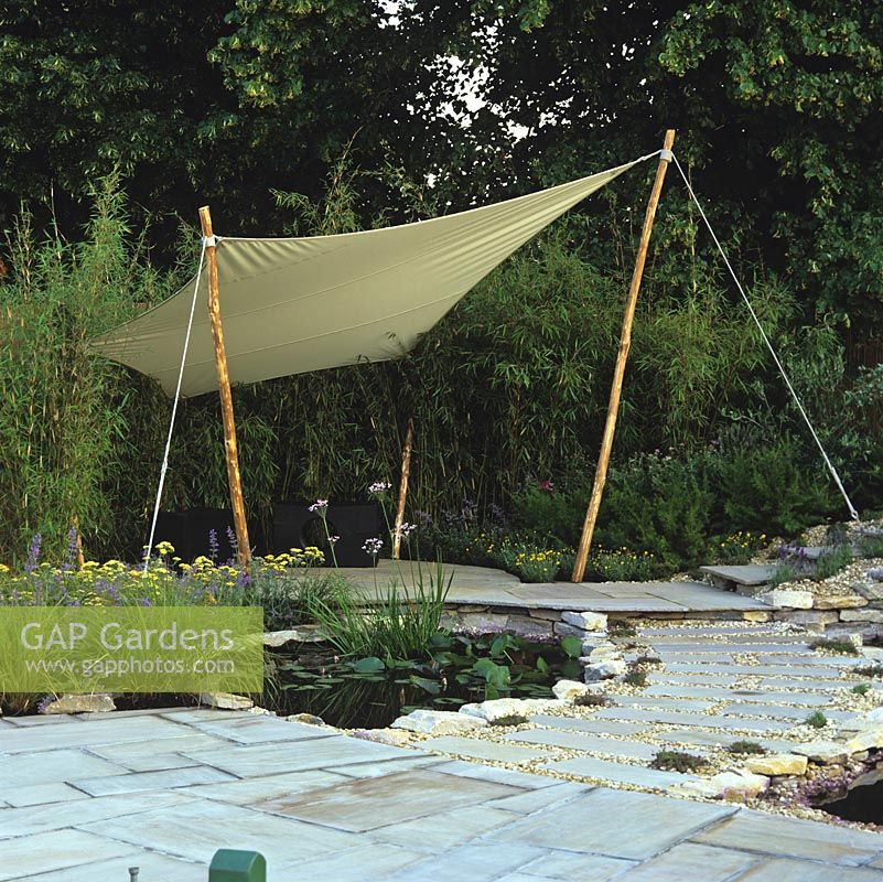 Sail awning on wooden poles by rustling bamboos enclose paved seating area by lily pond, edged in stepping stones set in chippings, paving, achillea, catmint and grasses.