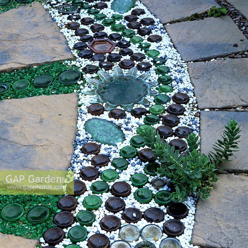 Patio design with paving slabs interspersed with lines of sliced bottoms from glass beer bottles or dishes, set in sand topped with green glass or white stone chippings.
