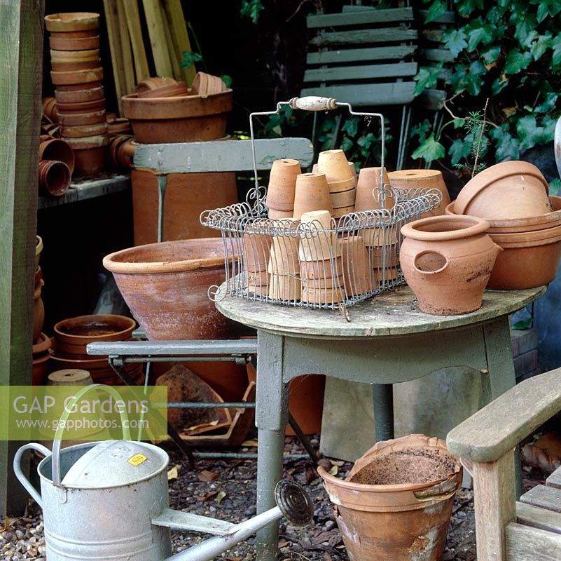 Collection of terracotta flower pots in a wire basket on table, watering can and old chair in working area.