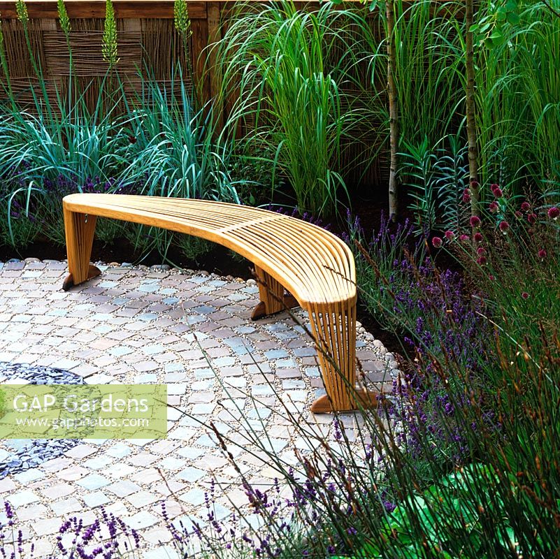 Organic shapes and sculptural planting. Swirling granite setts. Plants: restios, grasses, allium, lavender. Graceful wood bench. Woven willow panels at back.