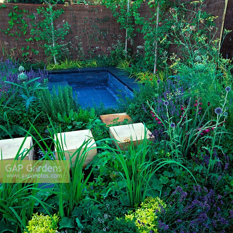 Wood oak cubes form stepping stones through planting and across small wildlife pool beside sunken, slate patio with wall seats for relaxing amongst aromatic plants.