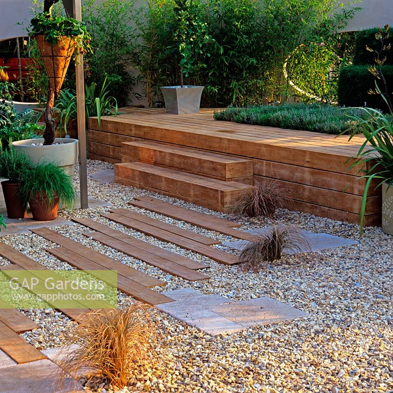 Wooden planks laid in gravel form path going up steps to raised wooden deck with inset bed of aromatic thyme. Behind, oval moon gate in wall between yew 