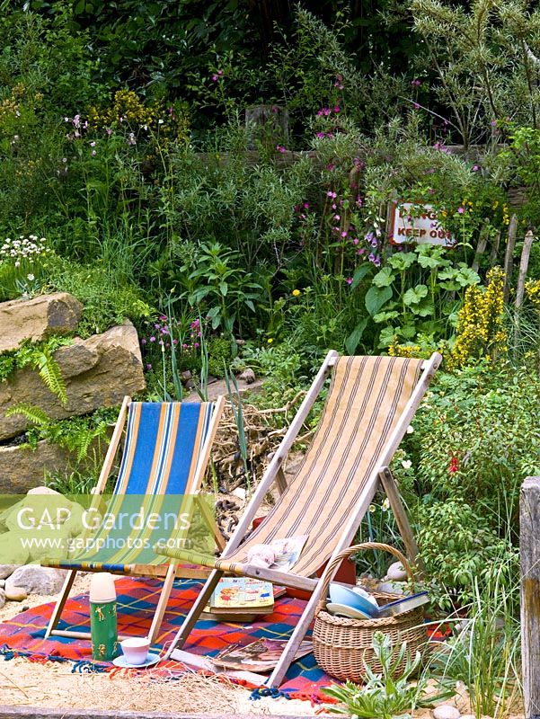 Seaside-themed garden with boulders retaining a bank of wildflowers, deckchairs and picnic basket on rug on sand.