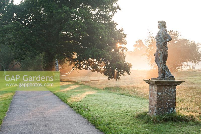 Statues bathed in rays of the rising sun, on the edge of the south lawn with ha ha and fields behind. Forde Abbey, nr Chard, Dorset, UK