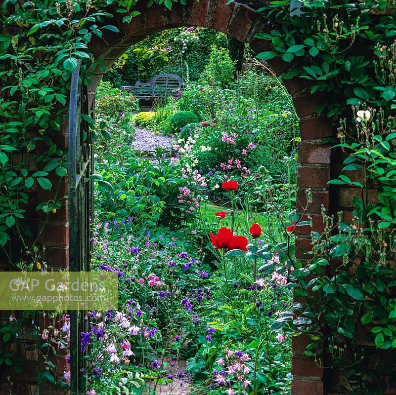 View through gate to Lutyens-style bench in herb garden. Beds of poppy, aquilegia, clematis, rose, hardy geranium and herbs punctuated by paths and box balls.