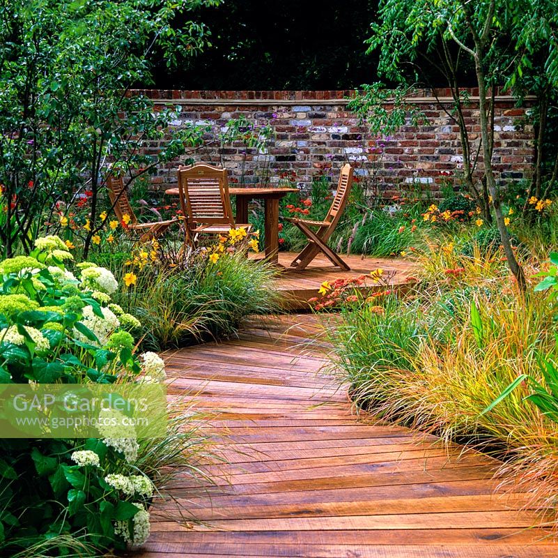 Seating area with table and chairs, on raised deck beneath canopy of birches. Curving timber path edged in grasses, hydrangea, daylily and achillea.
