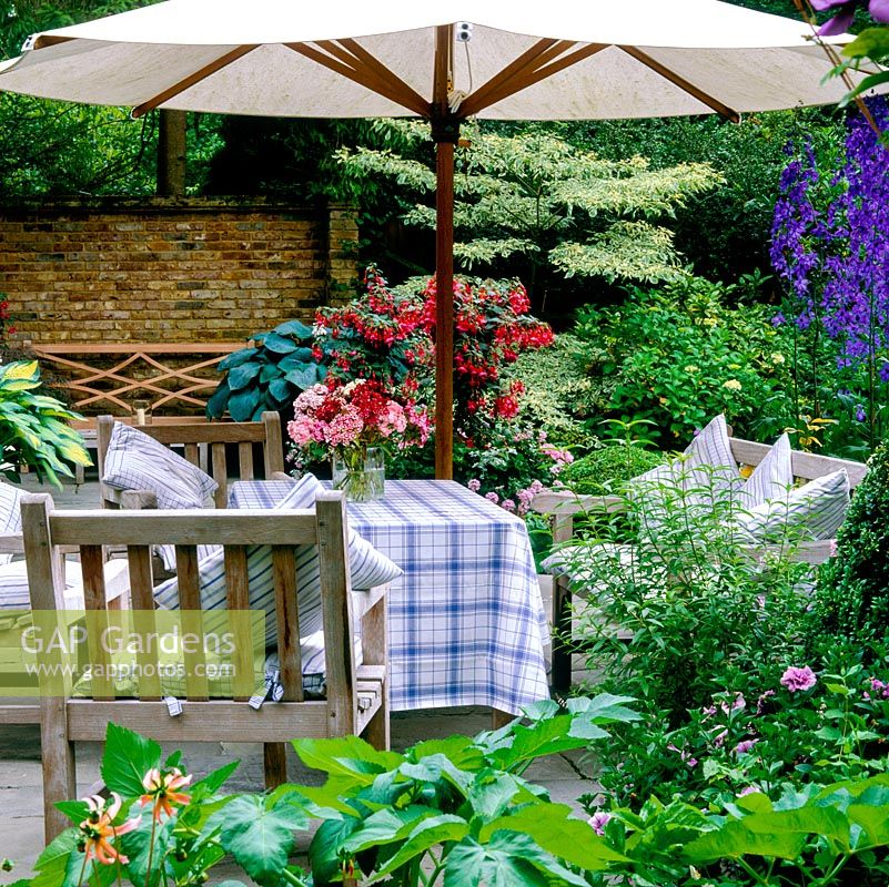 Stone terrace with pots of box, fuchsia, geranium, hosta, acer. Edged in beds of delphinium and cornus. Parasol shades wooden dining table, chairs, cushions. Vase of flowers.
