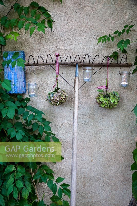 Garden rake decorated with hydrangea flowerheads and tealight candles in jars against a wall