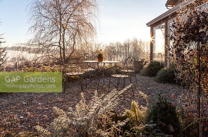 Conservatory, gravel terrace with metal garden furniture and mixed borders with weeping willow in the background - December, Mas de Bety, France