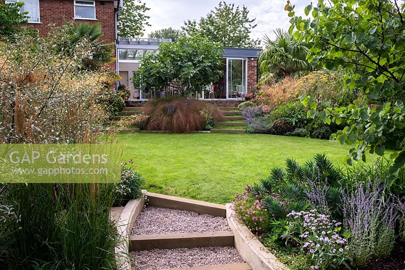 Gravel steps leading onto an oval shaped lawn in a split level terraced garden. In the foreground Calamagrostis 'Karl Foerster', Euphorbia, Verbena.