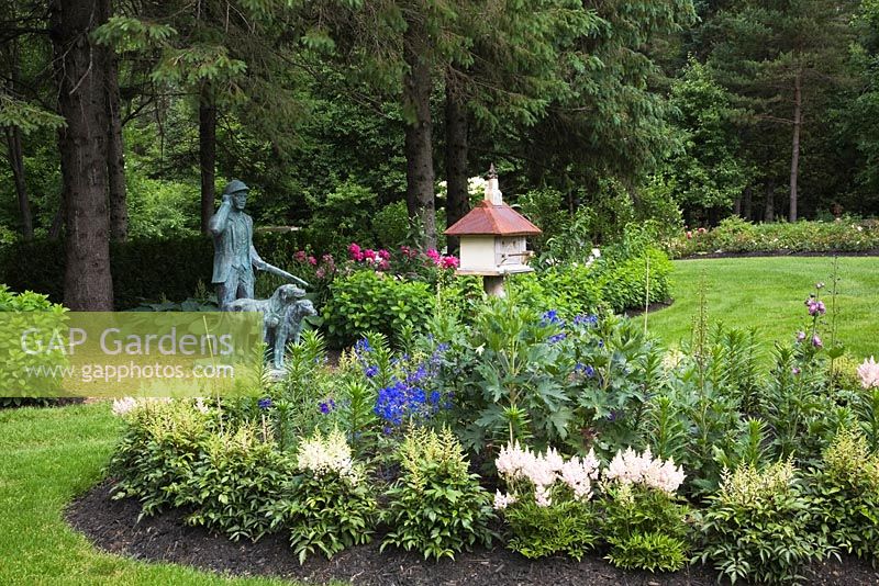 Statue of a hunter with his dogs next to border with wood and metal birdhouse and planted with pink and white Astilbe flowers in front yard country garden in summer, Quebec, Canada