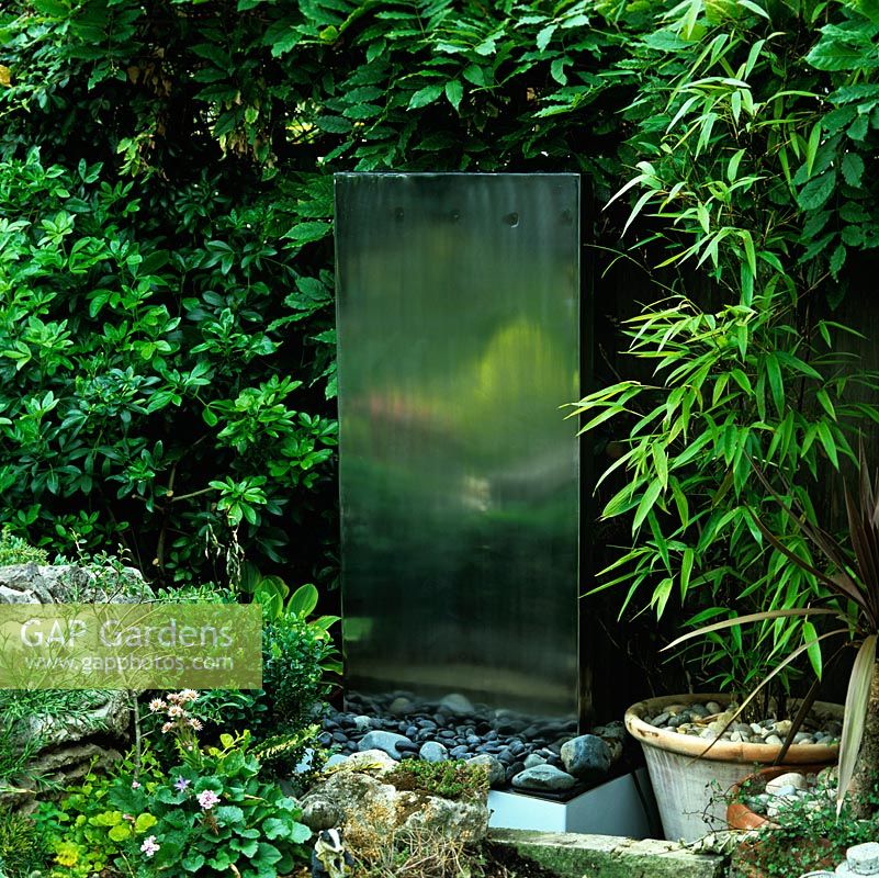 Stainless steel block water feature, set on bed of pebbles, dimly reflects rockery beyond.