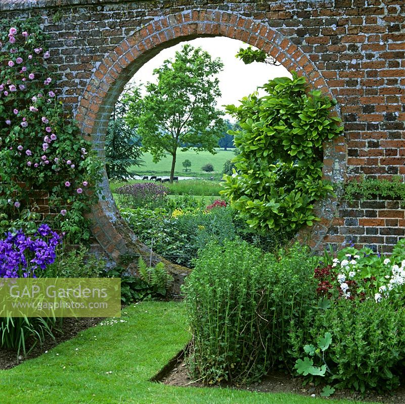 Circular moongate in old brick wall, flanked by roses, magnolia, iris and aquilegia, frames view of parkland, cows and distant countryside beyond.