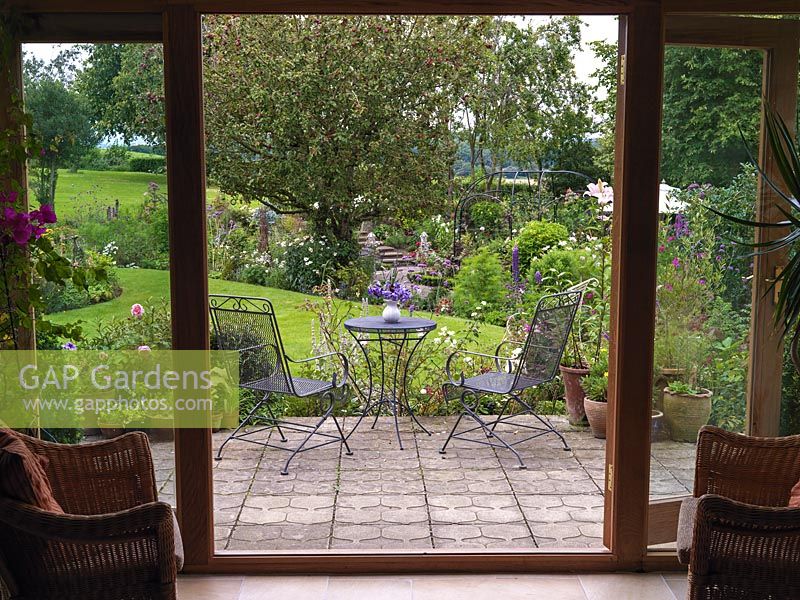 Patio doors frames view of terrace and country garden beyond with beds of lilies, roses, cosmos, Verbena bonariensis, lupin, allium, buddleja and old apple tree.