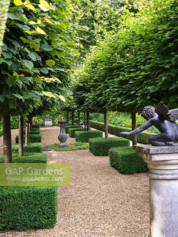 In secret, sunken formal garden, avenue of pleached limes, with their trunks encased in box squares. On pedestal, statue of Cupid. Central sundial.
