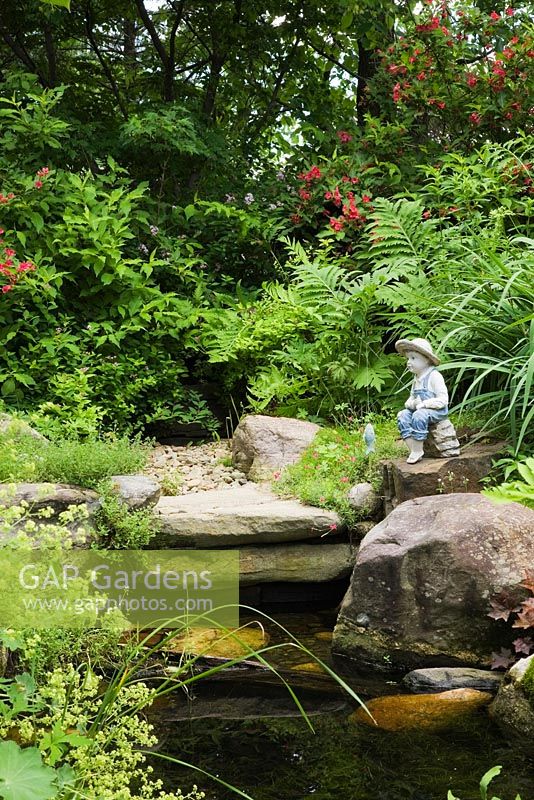 Sculpture of young boy fishing on the edge of a pond with Alchemilla mollis - Lady's mantle, Typha latifolia - Common Cattails and red flowering Weigela shrubs in backyard country garden in summer, Quebec, Canada