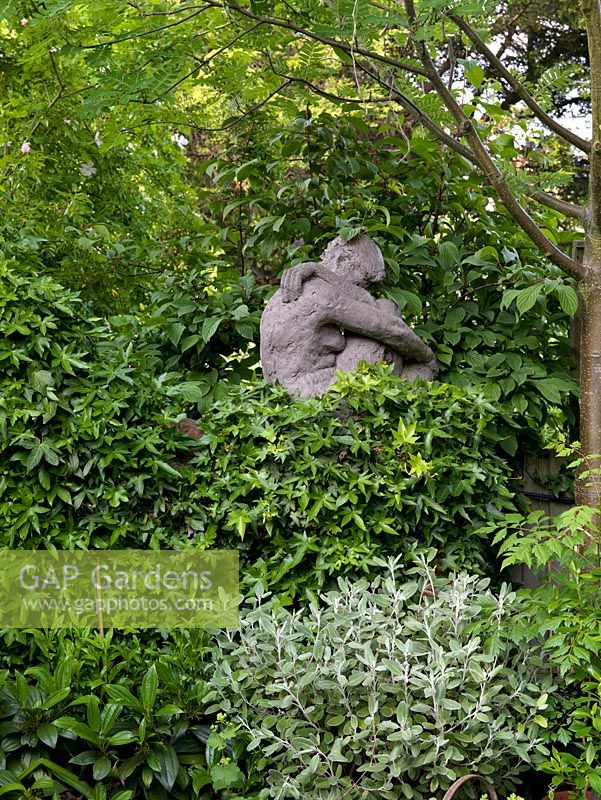 Tucked away under a sorbus tre, a sculpture of a woman and child sits on top of a wall covered in evergreen ivy.