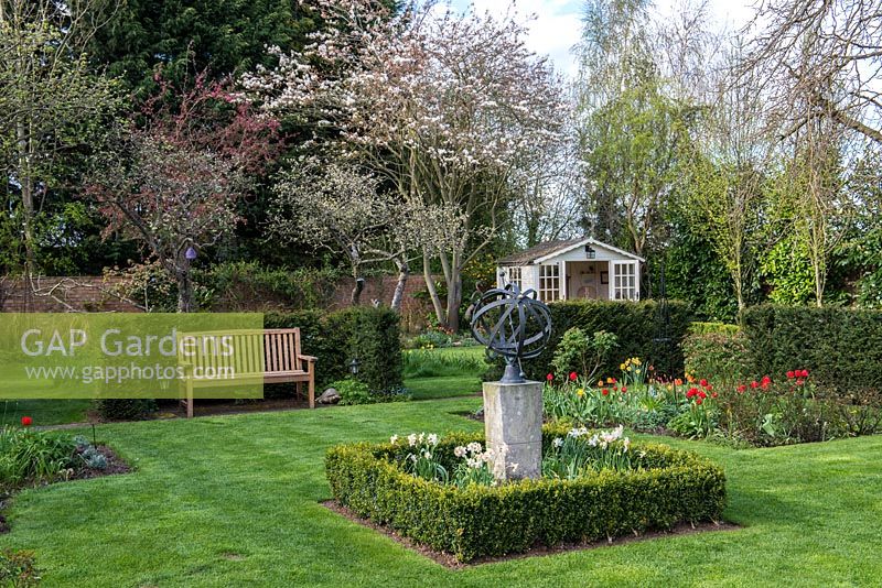 A formal grass parterre surrounded by yew - Taxus hedging. An Armillary sphere is set in boxed edged bed with Narcissus, the bed behind is planted with Tulips and roses. At the far end Amelanchier and Malus trees are in blossom.