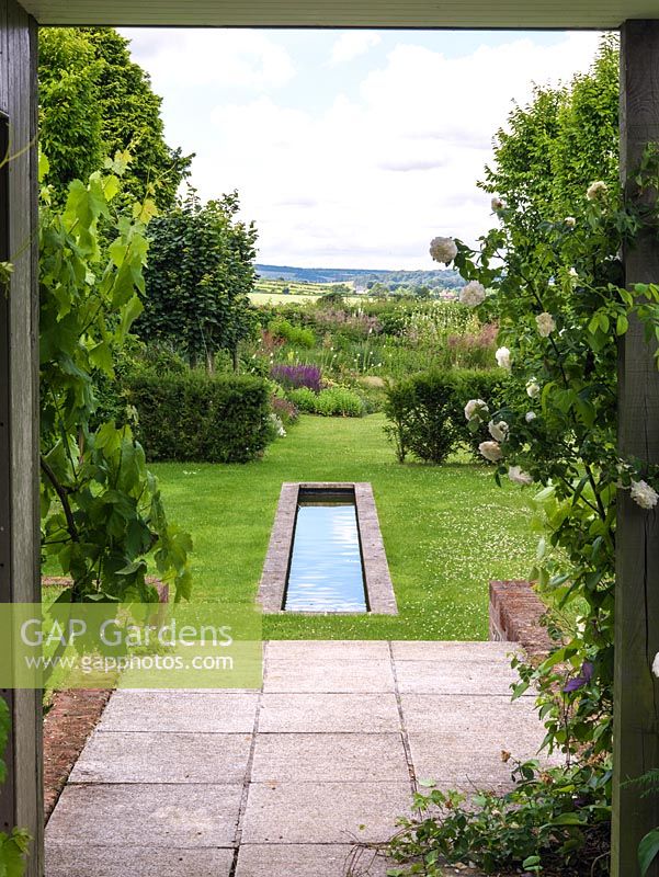Pergola clad in vine and Rosa 'New Dawn' frames view of rill. Beyond, naturalistic beds meld with countryside.
