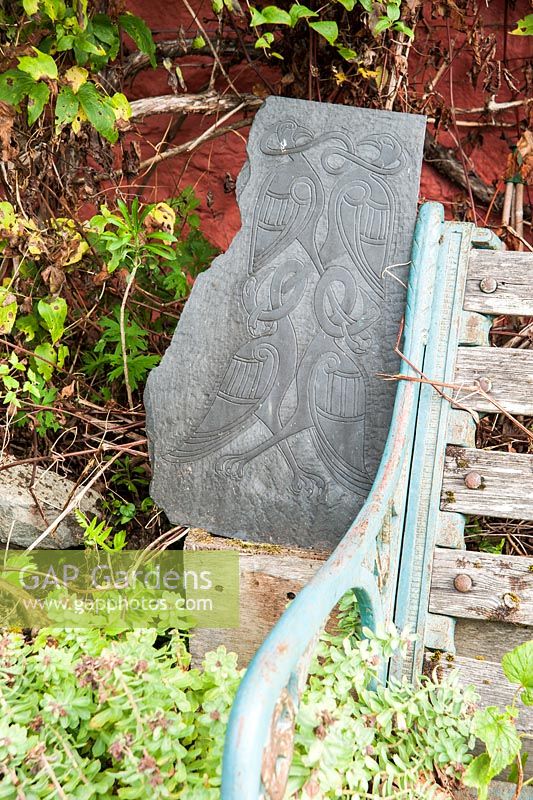 Decorative carved slate with birds beside bench in front garden. 