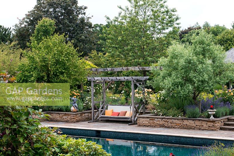 A view across a swimming pool to a swing seat between Cornus kousa 'Chinensis and Fraxinus angustifolia 'Raywood'.