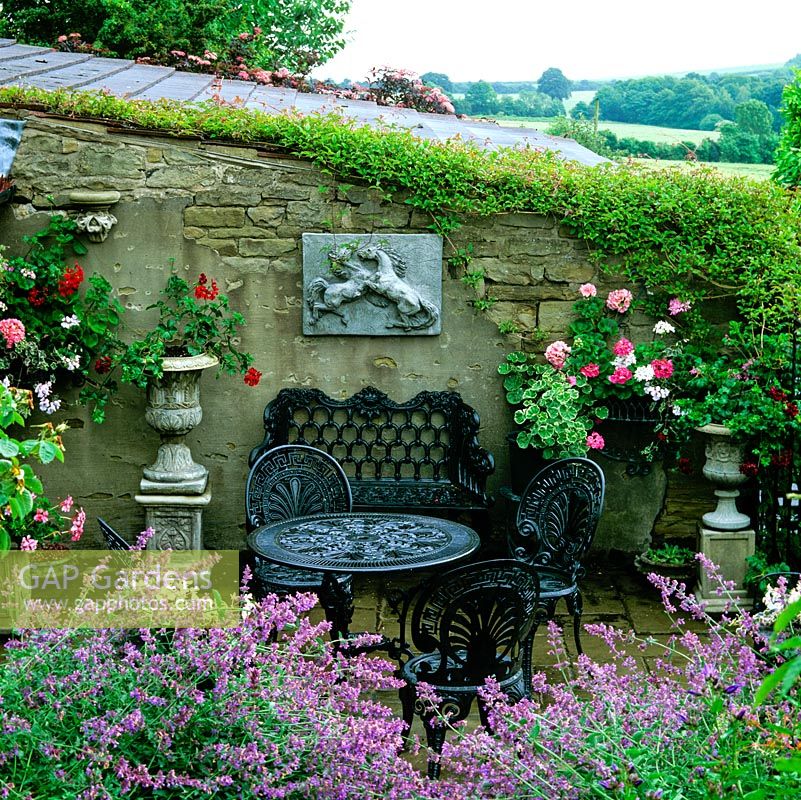 Sheltered by stone wall, sunken courtyard with wrought iron table, bench and chairs - wall with baskets and urns of geraniums. Edged in aromatic catmint. Behind, view of fields.