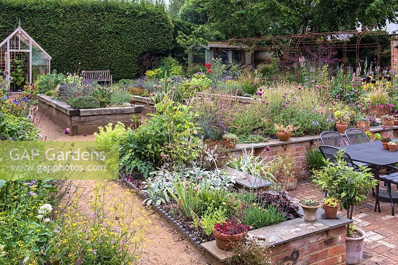 A split level, enclosed village garden with raised wooden beds planted largely with herbs, borders of late flowering summer perennials, a sunken terrace, pots of succulents and ornamental grasses, a wrought iron pergola and greenhouse.