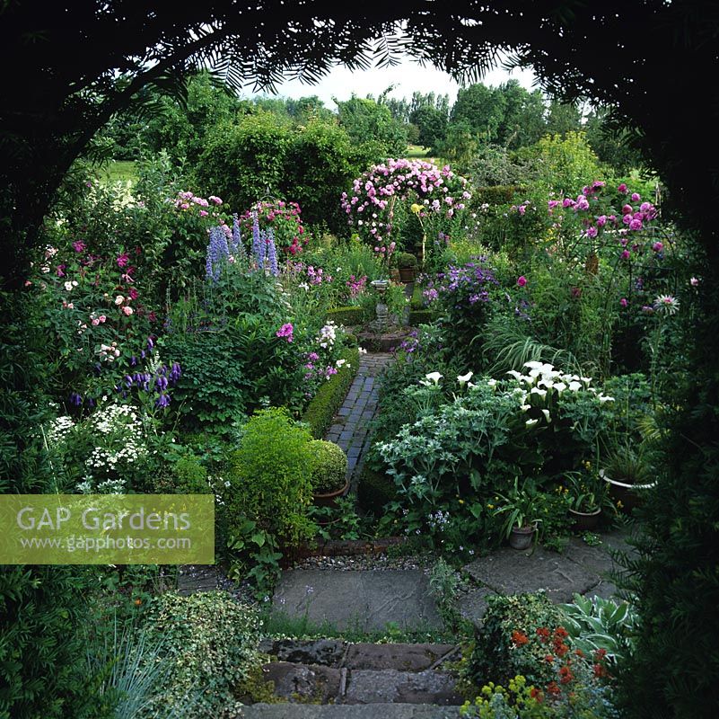 Yew arch leads to box parterre and sundial surrounded by beds of roses, Delphinium 'Alice Artindale', clematis, eryngium, lilies, pinks, scabious, aconitum, campanula and phlox.