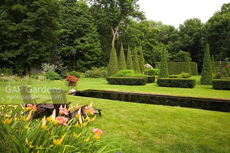 Orange Hemerocallis - Daylilies and pair of teak wood lounge chairs on manicured green grass lawn with Italian style mirror pool and clipped Thuja - Cedar trees and hedges in private backyard formal garden in summer