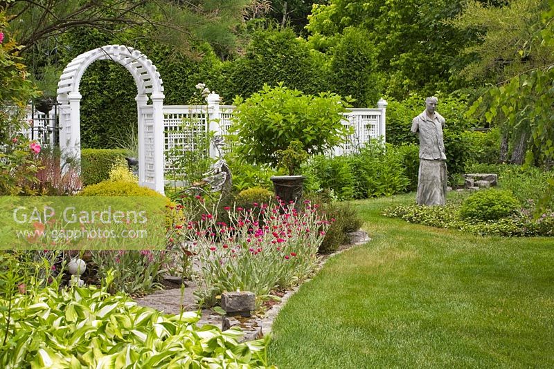 Manicured green grass lawn and border with Hosta plants, pink climbing Rosa - Rose bushes, purple Lychnis - Campion flowers, male concrete sculpture and white wooden arbour and lattice frame fence in private backyard formal garden in summer