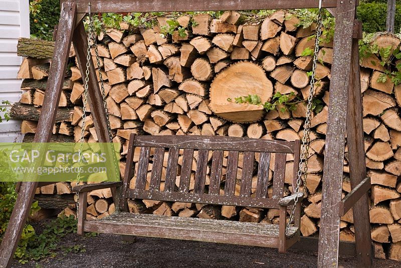 Brown painted wooden two seater swing in front of pile of stacked firewood in private backyard garden in autumn