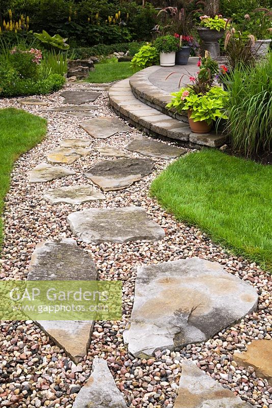 Flagstone and gravel path through manicured green grass lawn leading to raised brown paving stone patio with planters in residential backyard garden in summer