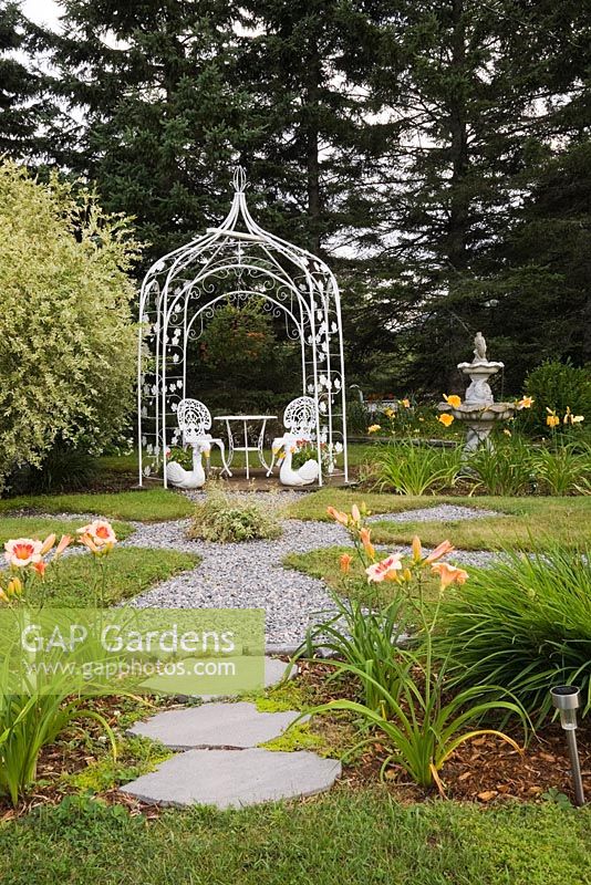 Flagstone and gravel path bordered by Hemerocallis 'Orange Crush' - Daylily flowers leading to white wrought iron pergola with a table and chairs in private backyard country garden in summer